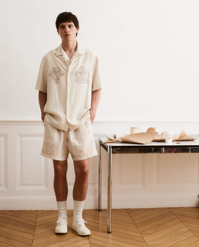 Model Joseph Signoret standing in an off-white embroidered short-sleeve shirt and matching shorts beside a metal table on parquet flooring.