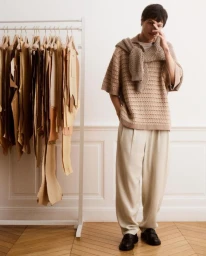 Model Joseph Signoret poses in a beige knit sweater and cream pleated trousers beside a clothing rack for Maison Kitsuné.
