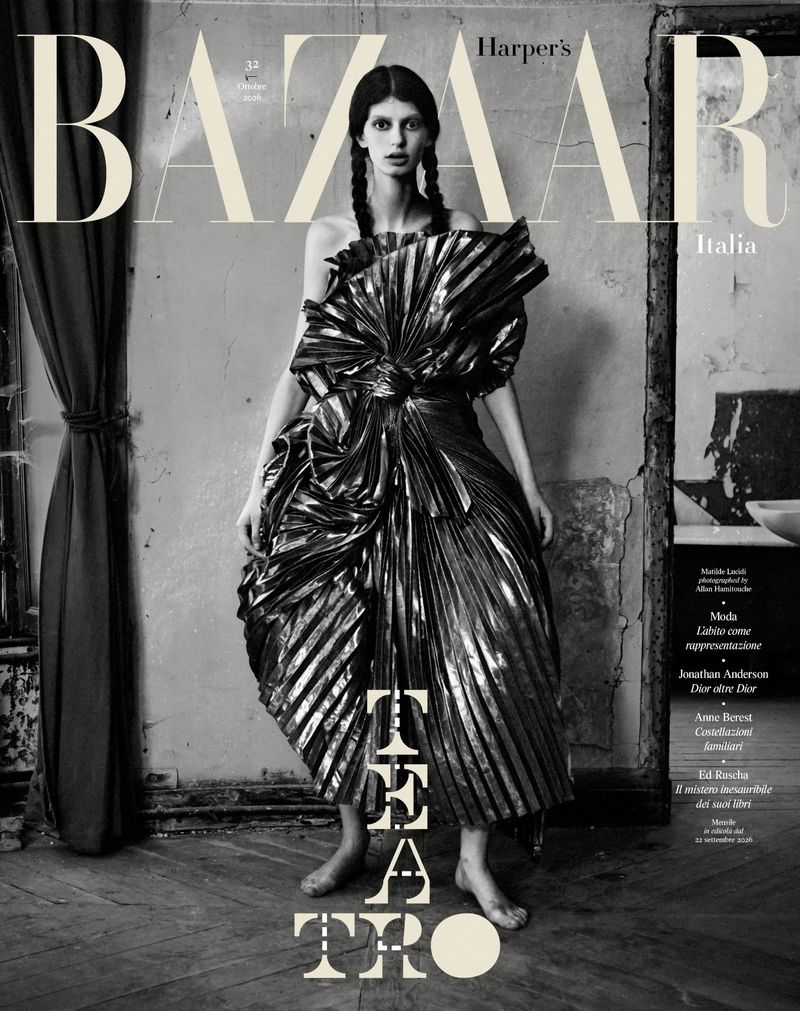 Monochrome magazine cover featuring a model in a voluminous metallic pleated gown standing in a weathered room.