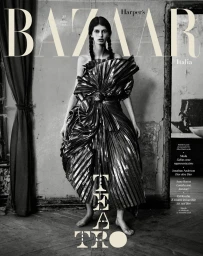Monochrome magazine cover featuring a model in a voluminous metallic pleated gown standing in a weathered room.