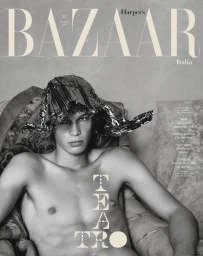 Shirtless male model wearing a metallic foil headpiece seated in an armchair for Harper's Bazaar Italia