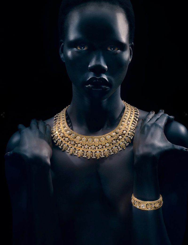 Model Naomi Apajok wearing an ornate gold necklace and bracelet against a dark background, photographed by Rob Rusling.