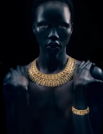 Model Naomi Apajok wearing an ornate gold necklace and bracelet against a dark background, photographed by Rob Rusling.