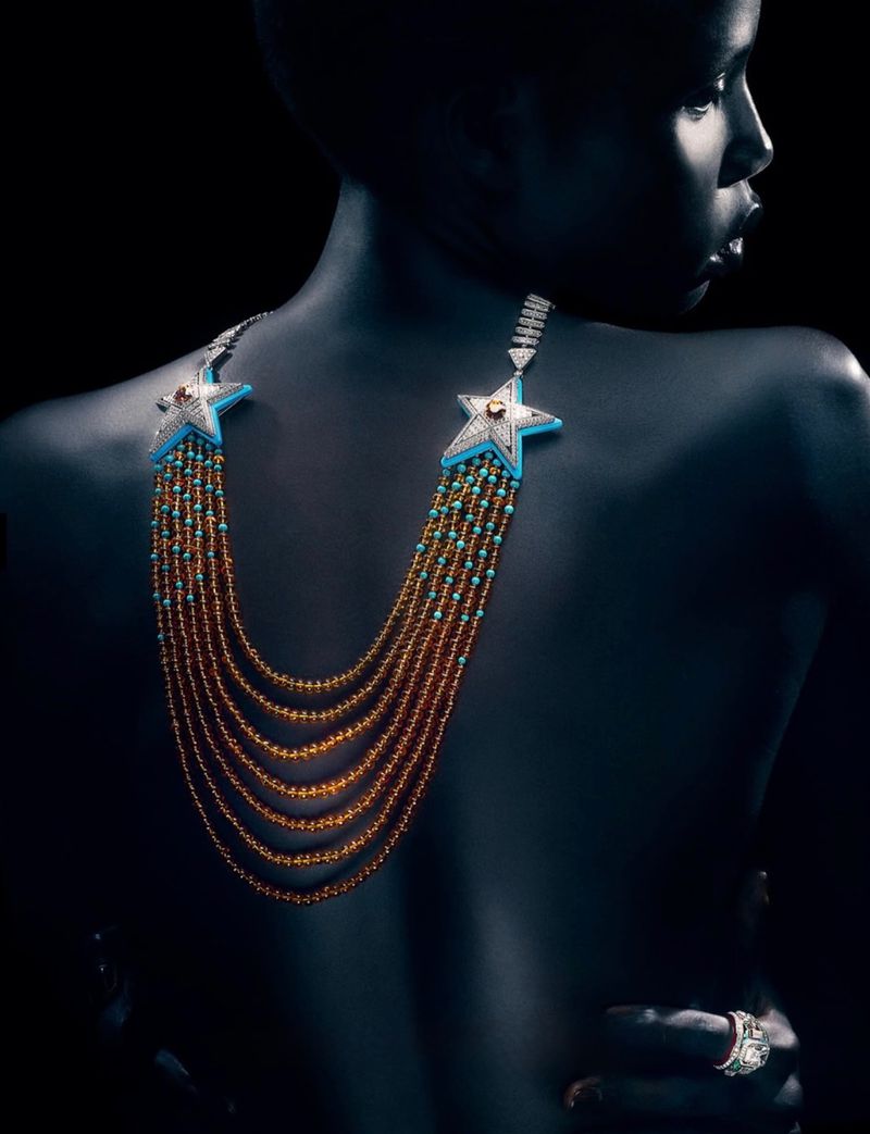 Model Naomi Apajok wearing an elaborate star-motif multi-strand beaded necklace against a dark background