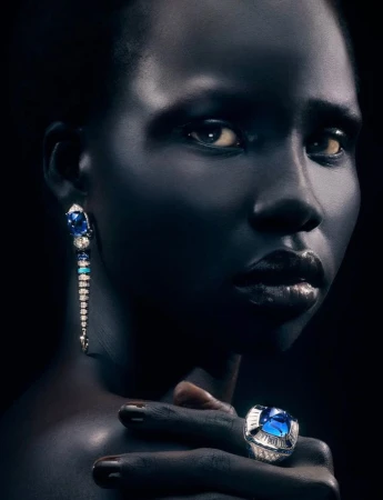 Close-up portrait of Naomi Apajok wearing a statement blue gemstone ring and dangling earrings against a dark background.