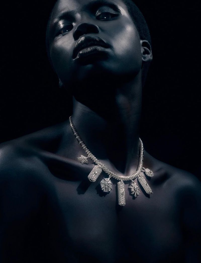 Close-up portrait of model Naomi Apajok wearing a diamond charm necklace against a dark background