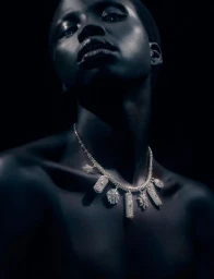 Close-up portrait of model Naomi Apajok wearing a diamond charm necklace against a dark background