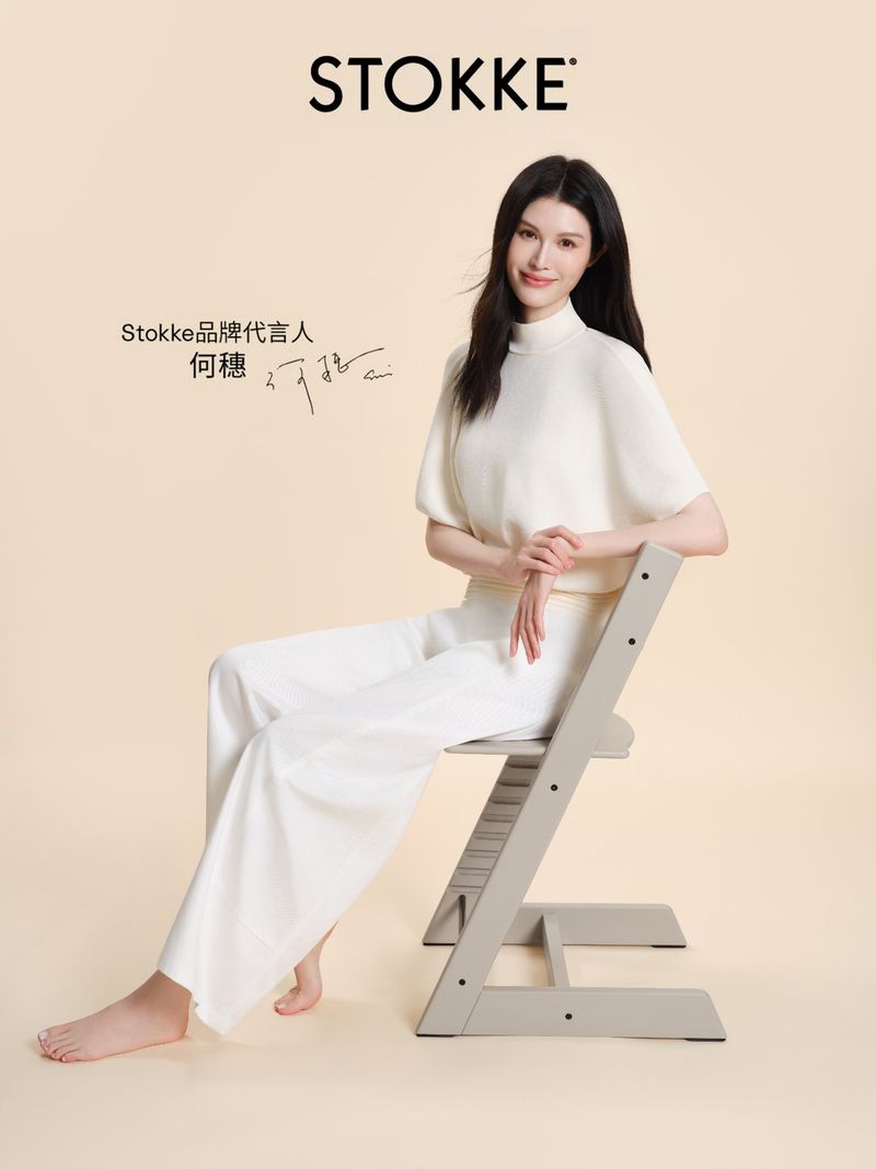 Model Sui He seated on a grey wooden high chair against a solid beige studio background, wearing white top and trousers.