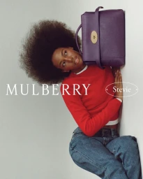 Model with voluminous afro hairstyle wearing a red sweater and holding a purple leather handbag against a light background.