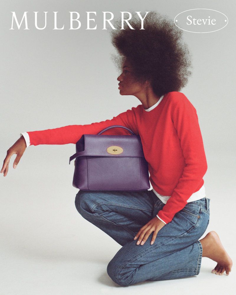 Model kneeling against a white backdrop holding a purple leather handbag, wearing a red sweater and blue jeans.
