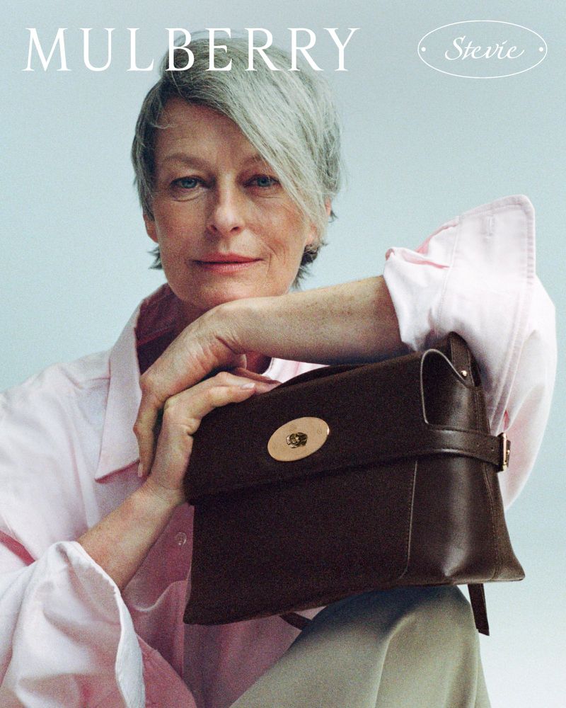 An older woman resting her chin on her hands while holding a dark brown leather handbag against a pale grey backdrop.