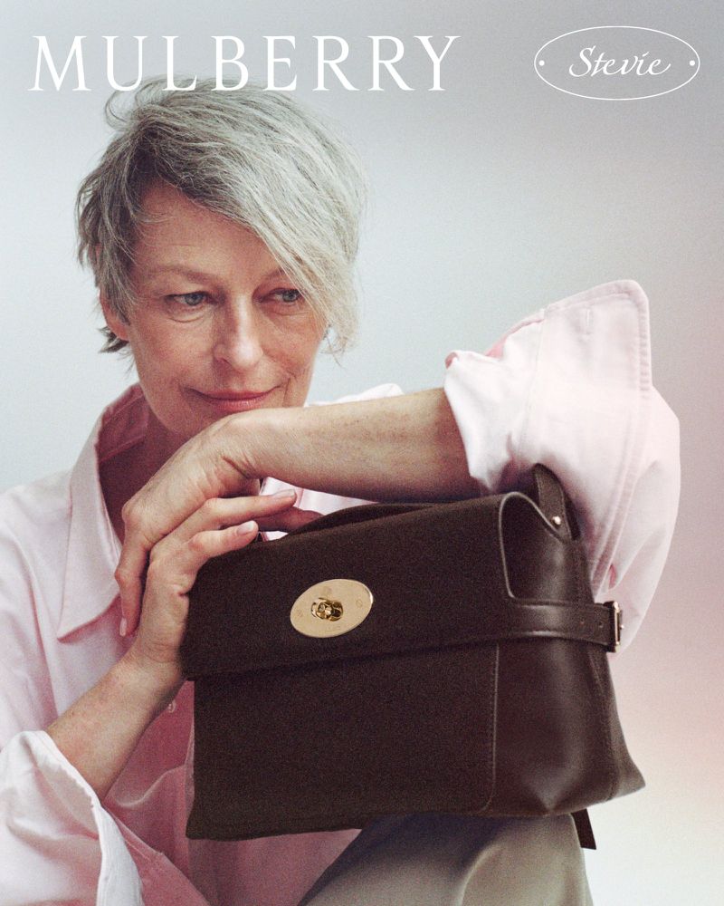 Model with short grey hair wearing a light pink shirt holding a dark brown leather handbag for Mulberry