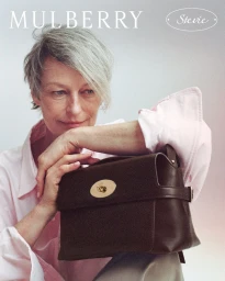 Model with short grey hair wearing a light pink shirt holding a dark brown leather handbag for Mulberry