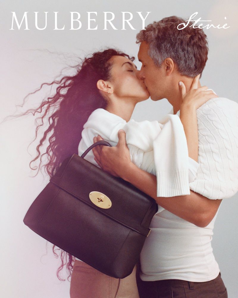 Man and woman embracing and kissing in front of a neutral studio backdrop with a dark leather handbag held in the foreground