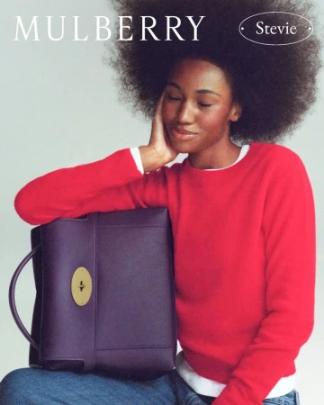 Black woman with afro wearing a red sweater holding a purple leather handbag against a plain grey background