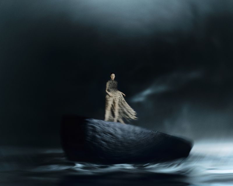 Woman in an airy dress stands on a rocky formation in a dark, atmospheric setting with motion blur, creating a dramatic visual effect.