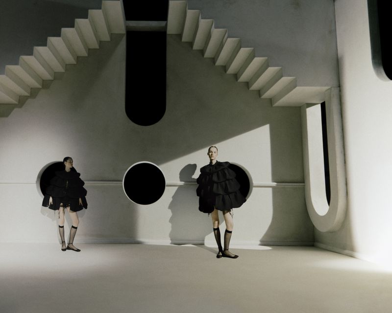 Two women pose in black ruffle dresses in an architectural space with stairs and circular openings against a light-grey backdrop.