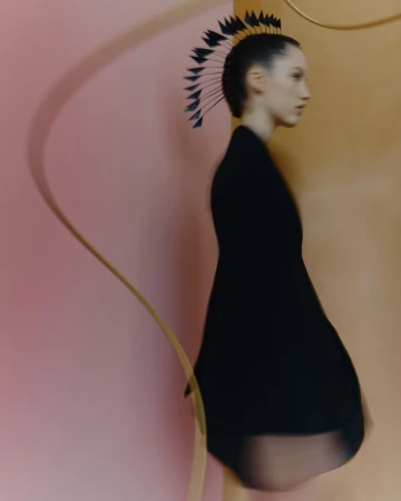 Profile shot of a woman with a dark feathered headpiece wearing a short black dress against peach and orange studio backdrop.