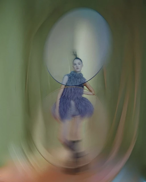Eye-level shot of a female fashion model posing indoors, wearing a feathered lavender dress, illuminated by soft light, through a round frame.
