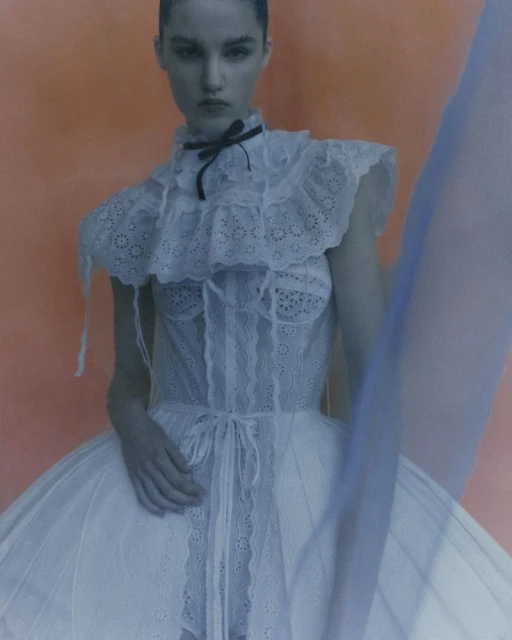 Fashion photo of a slender woman posing in a white dress with frills against a peach-colored background, creating a soft, elegant look.
