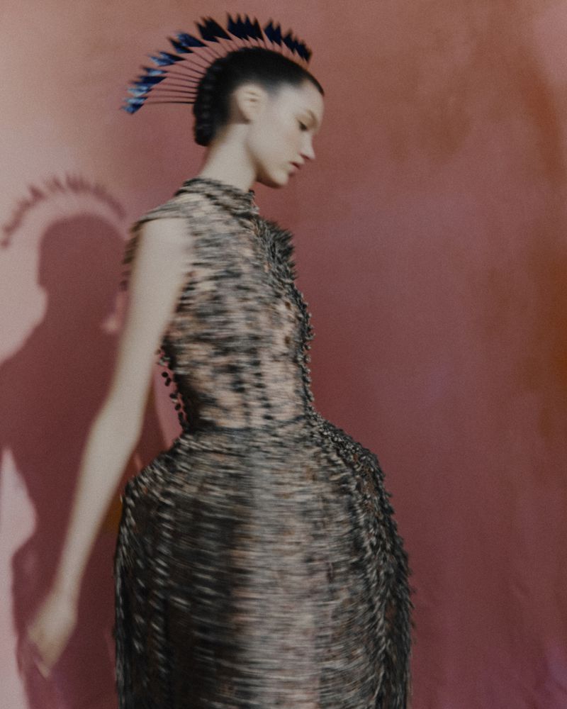 A stylish young woman with an avant-garde hairstyle and a textured dress poses in a fashion shot against a pink background.