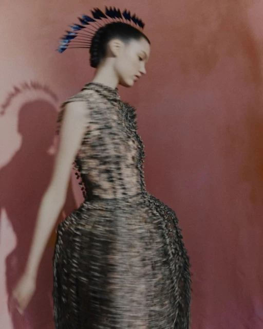 A stylish young woman with an avant-garde hairstyle and a textured dress poses in a fashion shot against a pink background.