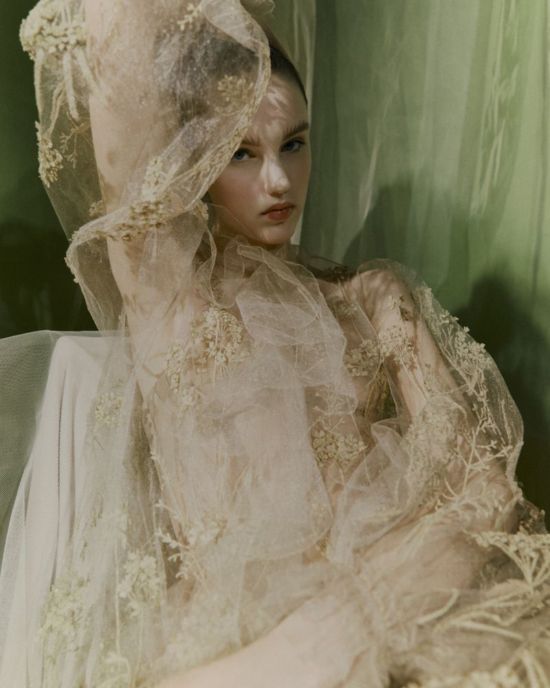 Woman with fair skin poses indoors draped in a sheer, embroidered fabric; soft focus and muted color palette.