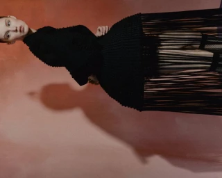 Model wearing a black structured textured knit dress with fringe details against a warm terracotta backdrop.