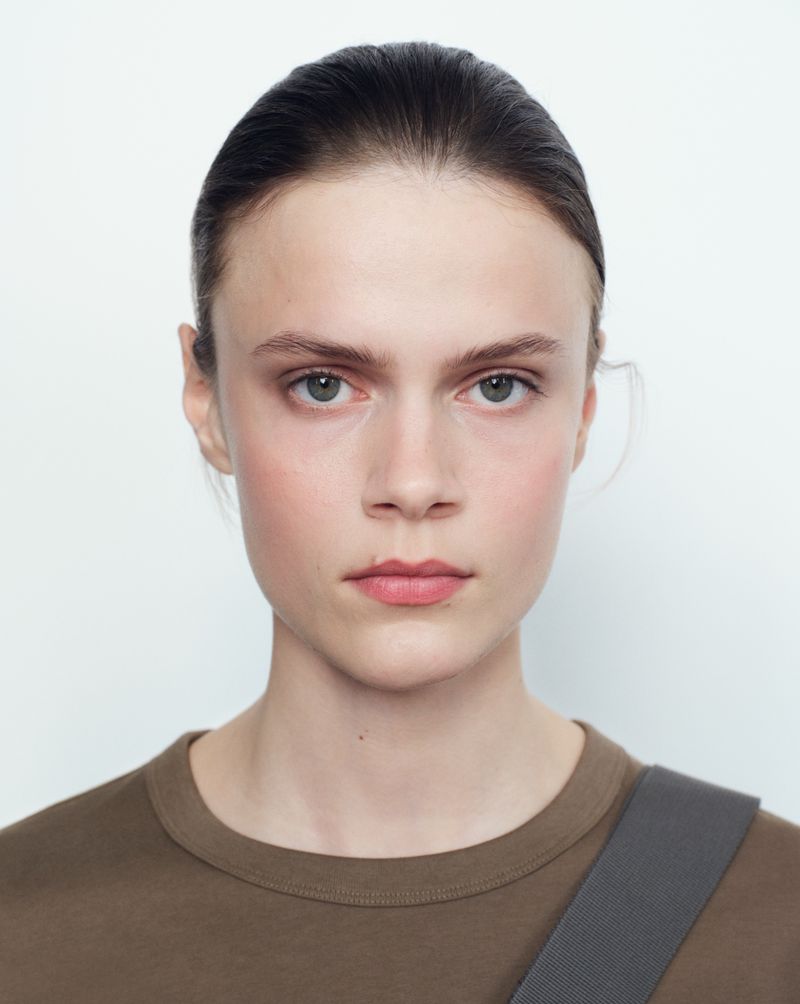 Close-up portrait of a model with dark pulled-back hair wearing a brown t-shirt against a plain white background