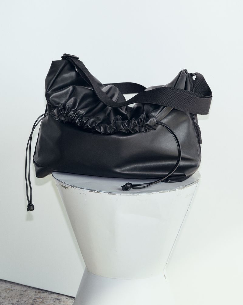 Black slouchy shoulder bag with drawstring details resting on a white sculptural pedestal against a plain background