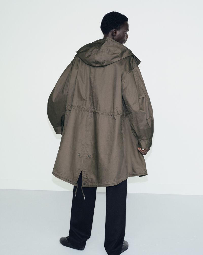 Model seen from behind wearing an olive green hooded parka and black trousers against a neutral white backdrop