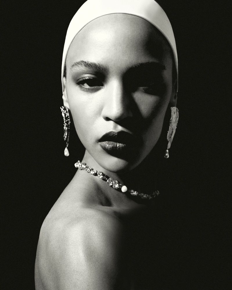 Close-up monochrome portrait of a model wearing a tight white cap, drop earrings, and a beaded necklace against a dark background.