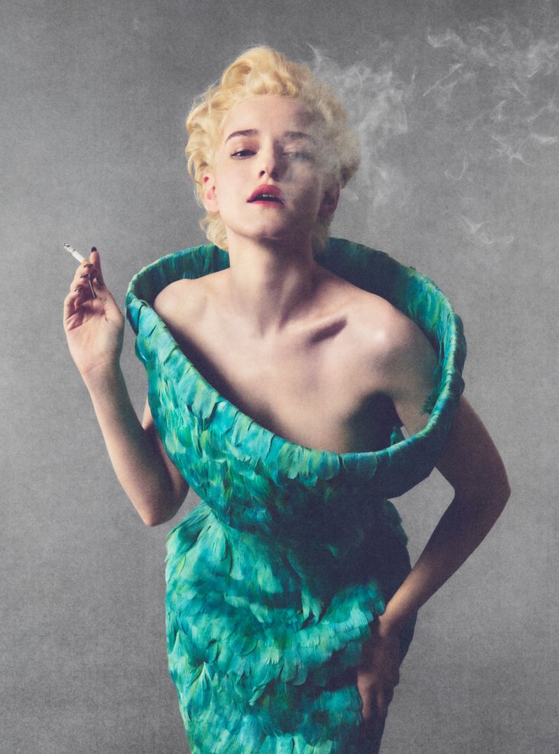 Julia Garner in a sculptural green feather dress holding a cigarette against a grey textured background for Elle U.S.