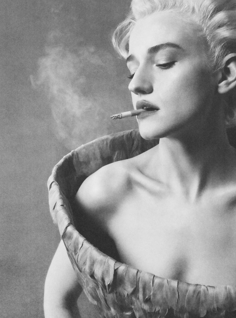 Julia Garner smoking a cigarette in a structured garment against a textured grey background.