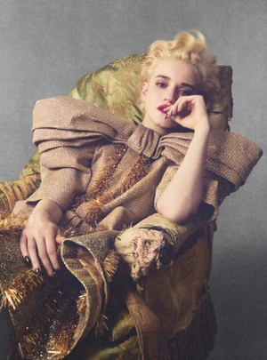 Blonde woman seated in an ornate patterned armchair wearing a textured structured garment with exaggerated shoulders.