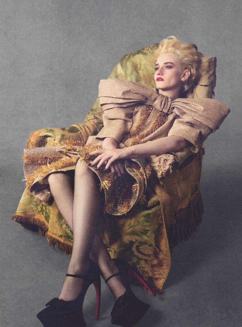Julia Garner seated in an ornate patterned armchair wearing a textured top with a large bow detail and black heels