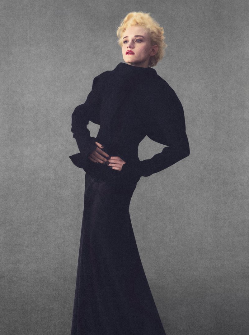 Julia Garner in a black high-collar jacket and long skirt against a grey textured studio backdrop for Elle U.S.