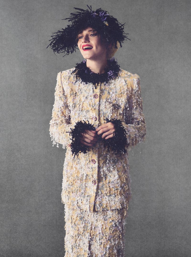 Julia Garner in a textured tweed jacket and feathered hat smiling against a grey textured background for Elle U.S.