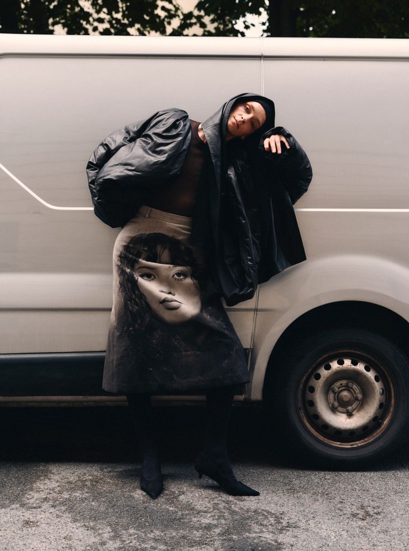 Model leaning against a white van wearing a black puffer jacket and printed midi skirt for Mixte