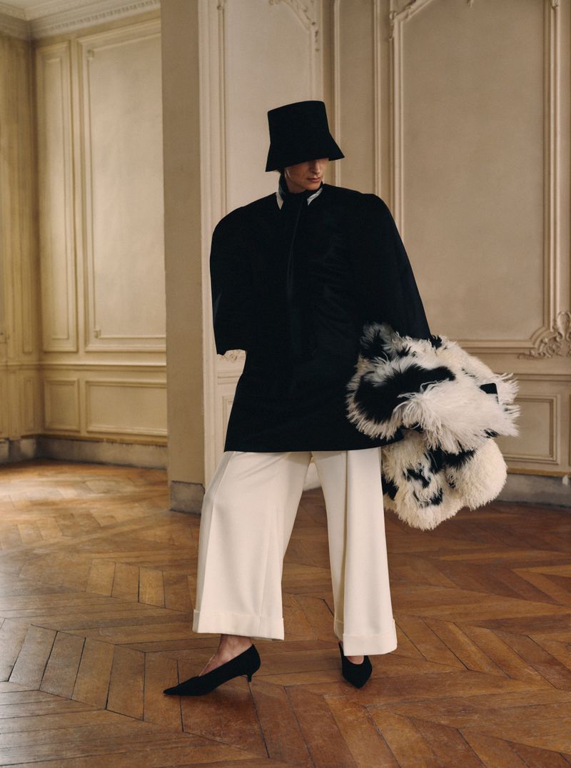 Model in black jacket, hat, white trousers, holding textured black and white garment in classical room