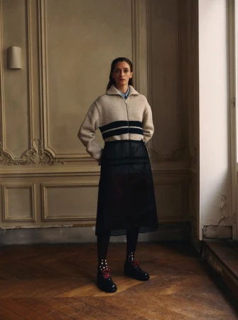 Model wearing a cream knit zip sweater and dark sheer skirt standing in a classic room with wood paneling.