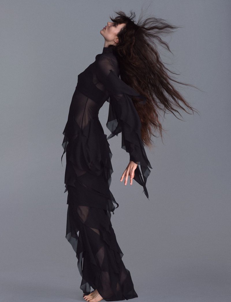 Grace Hartzel in a long sheer black ruffled dress with dark blowing hair against a grey seamless background