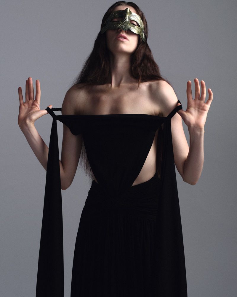 Model Grace Hartzel wearing a black dress and gold mask against a plain grey backdrop for Pop Magazine.
