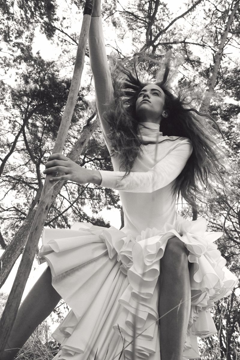 Monochrome fashion editorial image of Grace Hartzel wearing a white ruffled dress outdoors holding a wooden pole