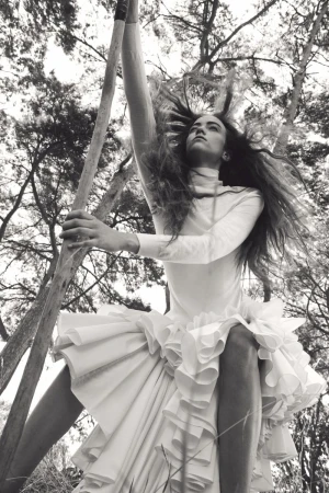 Monochrome fashion editorial image of Grace Hartzel wearing a white ruffled dress outdoors holding a wooden pole