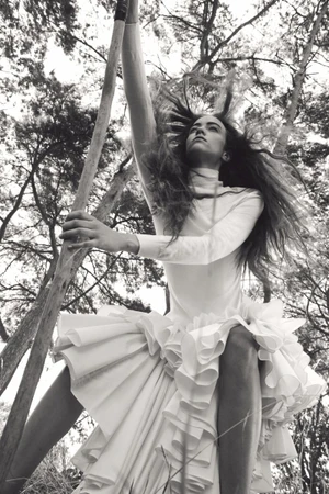 Monochrome fashion editorial image of Grace Hartzel wearing a white ruffled dress outdoors holding a wooden pole