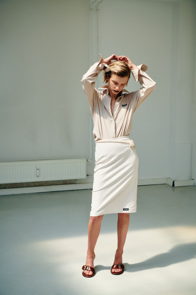 Model in cream blouse and white skirt with hands in hair against a white wall.