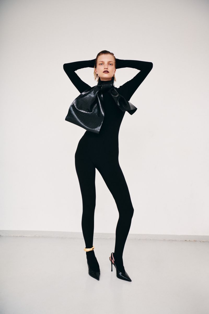 Model in black turtleneck catsuit wearing a large glossy leather bow against a plain white background