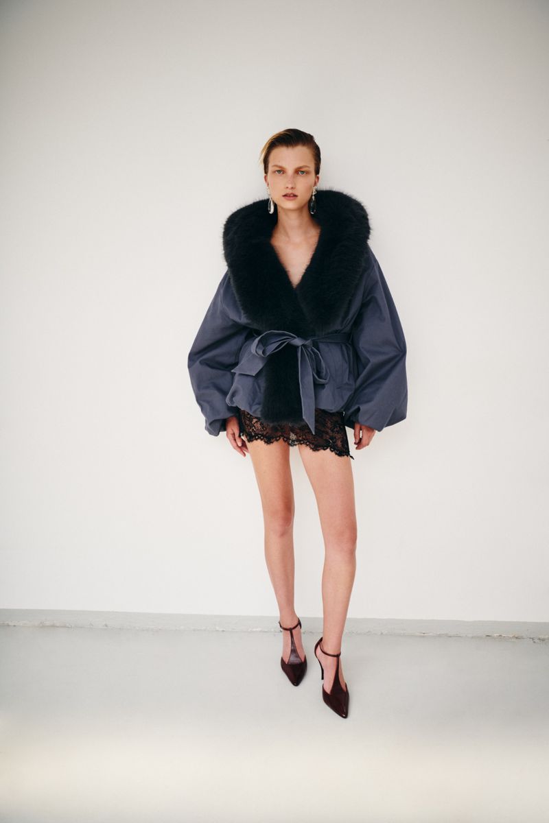 Model wearing a voluminous dark jacket with a fur collar and sheer lace skirt against a plain wall