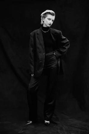 Model in black pinstripe oversized suit and dark turtleneck posing against dark textured backdrop